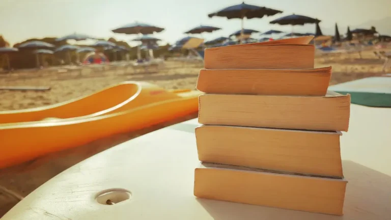 Pila di libri gialli e thriller appoggiati su una canoa in spiaggia, consigli di lettura per l'estate