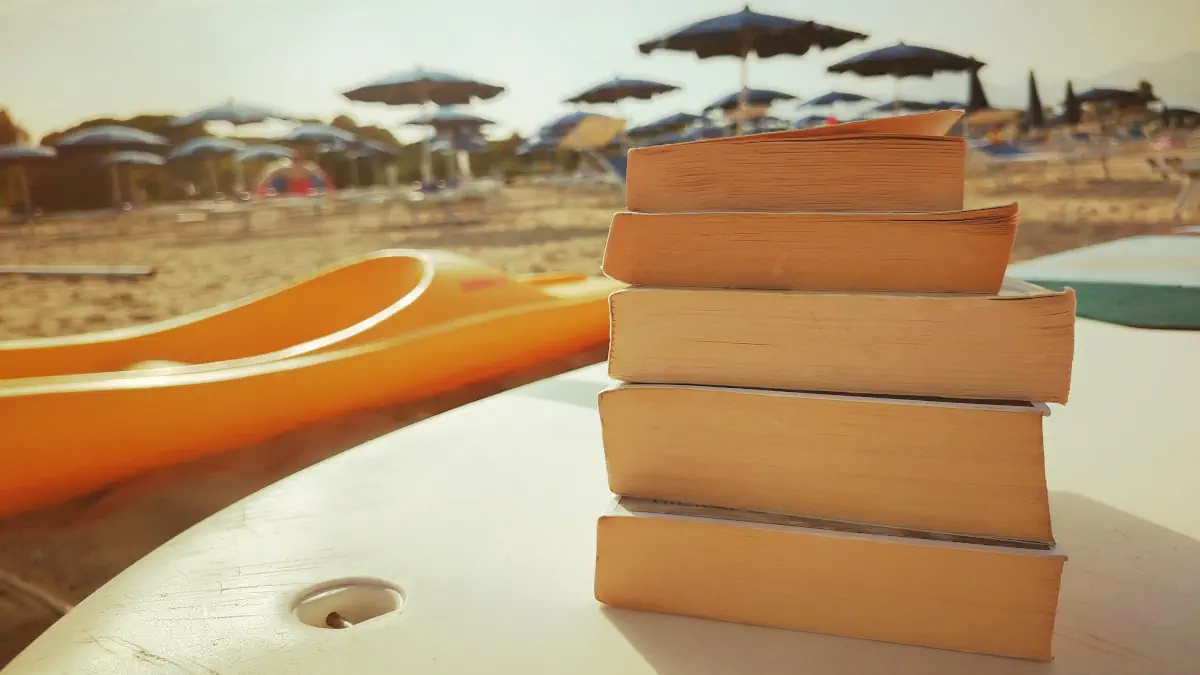 Pila di libri gialli e thriller appoggiati su una canoa in spiaggia, consigli di lettura per l'estate