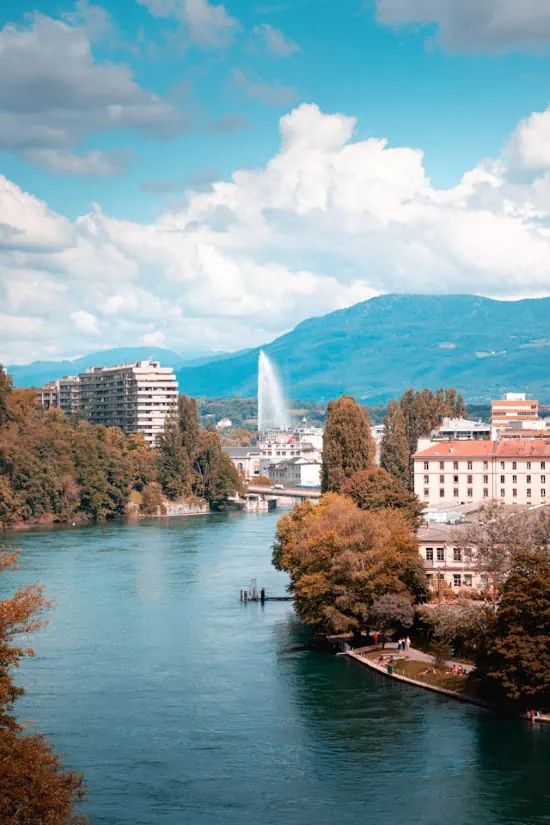 Scorcio panoramico della città di Ginevra con il fiume Rodano, alberi in primo piano e il famoso getto d'acqua Jet d'Eau sullo sfondo sotto un cielo nuvoloso. 