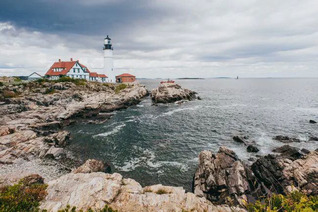 Il faro bianco Portland Head Light situato sulla costa rocciosa del Maine, con onde del mare increspate in primo piano sotto un cielo nuvoloso.

